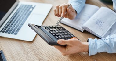 Cropped view of professional serious finance manager, holding calculator in hands, checking company month's profits, looking through details on laptop, writing down information for meeting with director